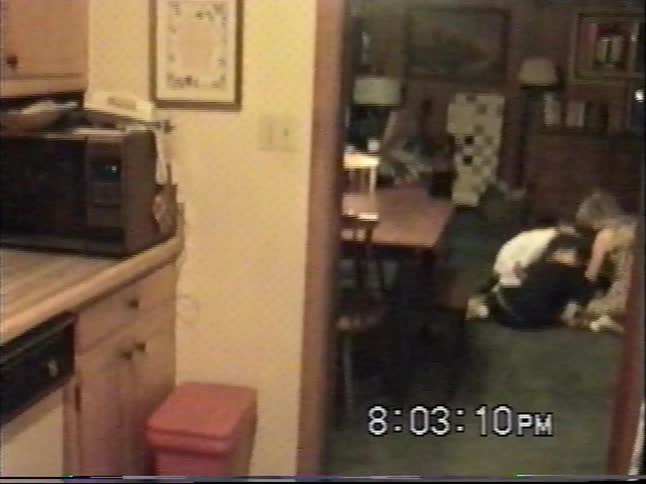 A grainy VHS home video from the 1990s shows a mother and her two sons lovingly petting their big black dog on the living room floor