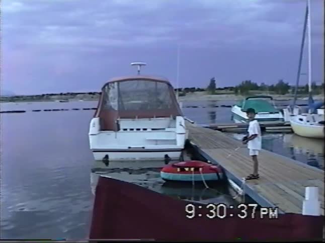 VHS footage captures a serene pink sunset over a marina where two young boys are fishing from the wooden dock