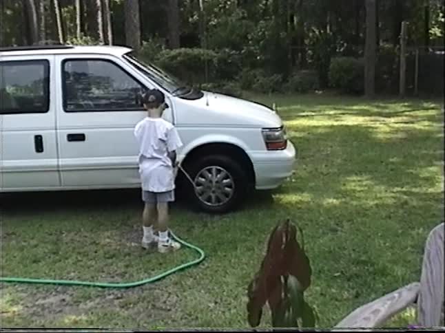 A young boy helps with chores by washing the family's white minivan in the grassy yard, vintage 1997 home video footage