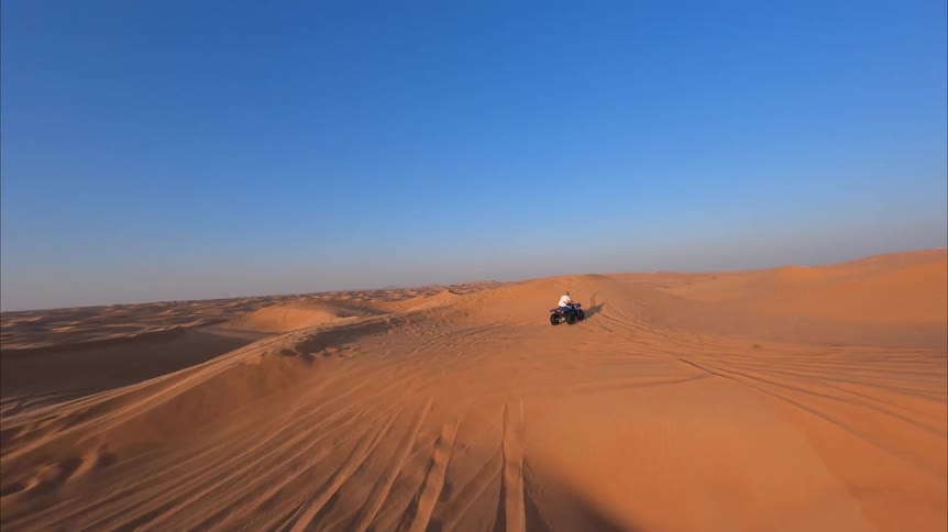 A first-person view follows a quad bike as it skillfully navigates the vast, rolling orange sand dunes of a desert under a clear blue sky