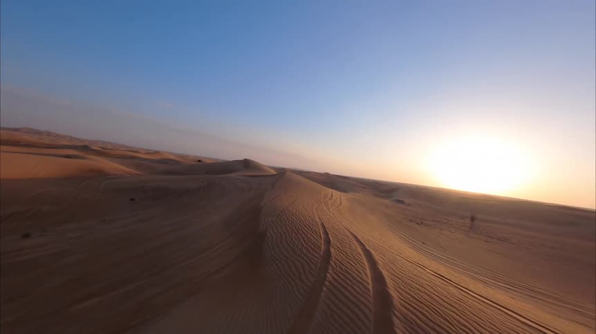 An FPV drone soars low and fast over the expansive golden sand dunes during a beautiful sunrise
