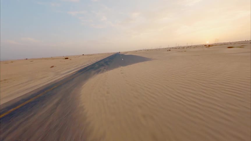 A dramatic FPV drone shot speeds down a deserted road partially covered by windswept sand dunes in the desert at sunrise