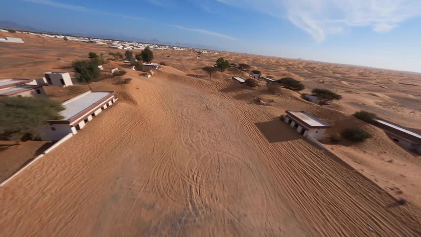Dynamic FPV drone footage soars over a deserted village half-buried in the golden desert sand under a bright blue sky
