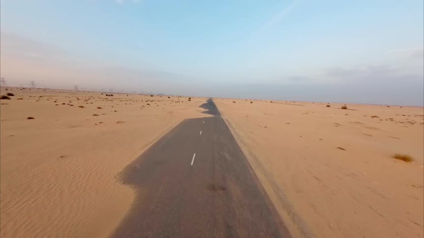 An FPV drone soars over a long, empty paved road that is being swallowed by the vast, golden desert sand dunes