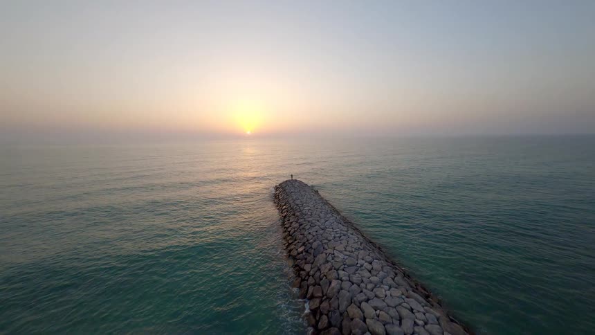 An aerial shot slowly approaches a solitary man standing on a rocky jetty, admiring the tranquil sunrise over the vast ocean