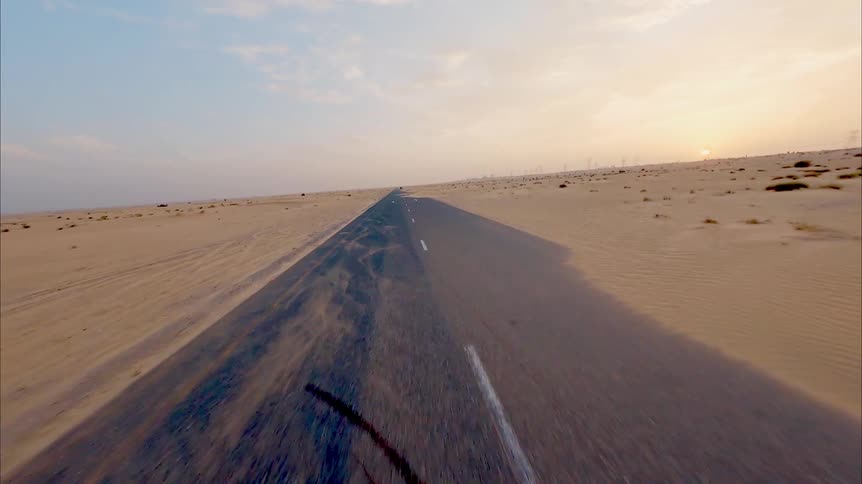 First-person view flying down a long, empty road cutting through a vast desert as sand blows across the asphalt at sunset
