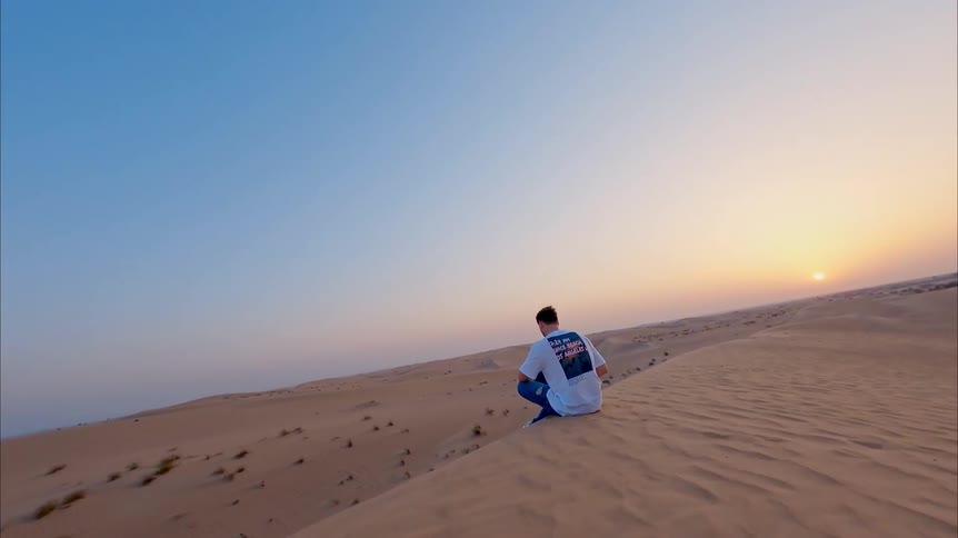Aerial shot of a man working remotely on his laptop from the top of a sand dune in a vast desert during a beautiful sunset