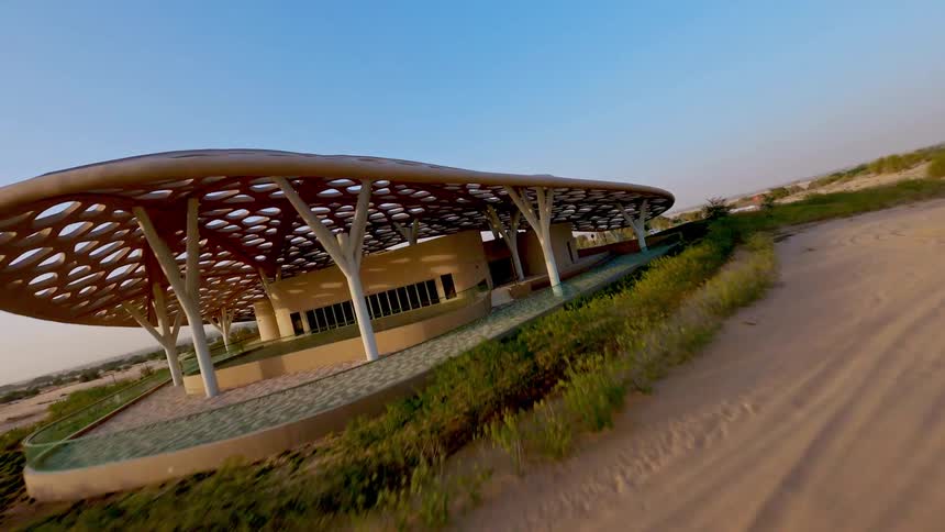 An FPV drone glides low over expansive desert sand dunes before rising over a stunning modern architectural building at sunset