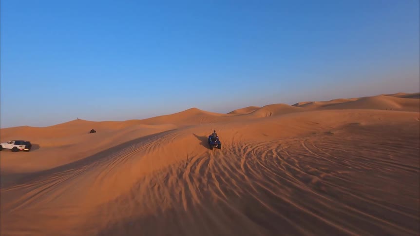 An FPV drone follows a person riding a quad bike through vast, rolling desert sand dunes during a beautiful golden sunset