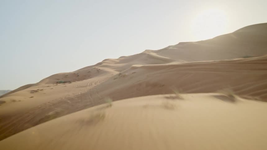 A drone's perspective flies low over the golden sand dunes of a vast desert with distant mountains and a bright sun