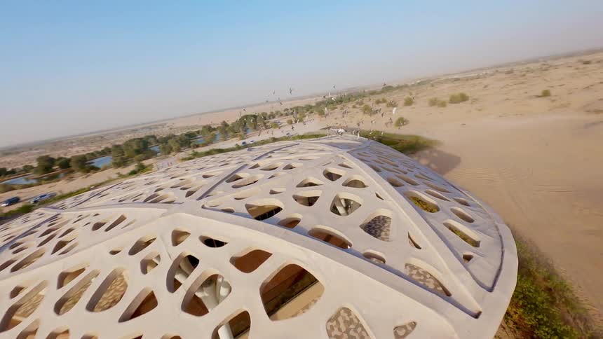 An aerial FPV drone view shows birds flying off a futuristic building in the Al-Wathba Desert and gliding over the sand dunes