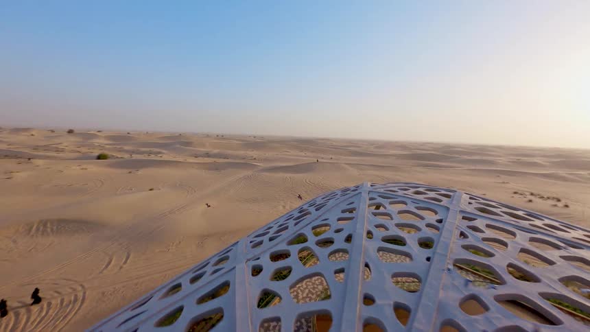 Sweeping aerial footage reveals a futuristic architectural masterpiece with a unique lattice roof, set against the backdrop of endless sand dunes and a desert oasis at sunset