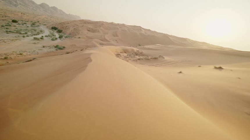 An FPV drone soars low over the vast, rolling golden sand dunes of a desert at sunset, revealing mountains in the distance