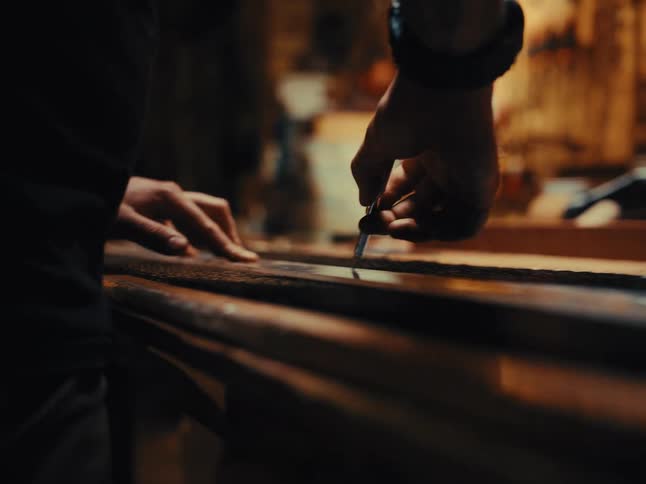 A craftsman with tattoos on his arm carefully uses a ruler to mark a straight line on a textured material in a workshop