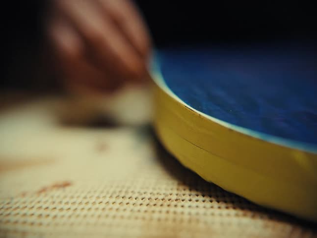 A close-up shot of an artist's hands carefully removing yellow masking tape from the edge of a blue circular resin art piece