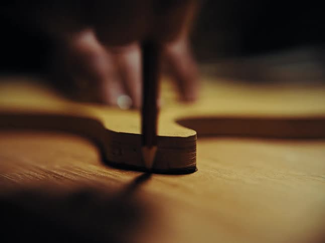 An artisan carefully traces the outline of a wooden guitar template onto a fresh plank with a pencil