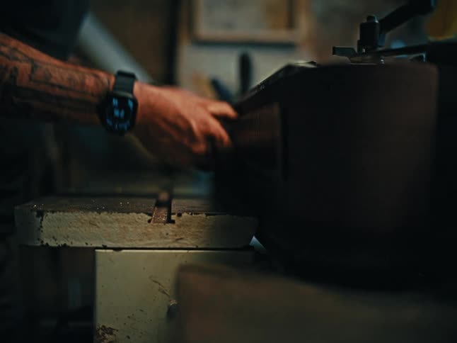 A craftsman with a tattooed arm uses a belt sander to smooth a piece of wood in a dimly lit workshop