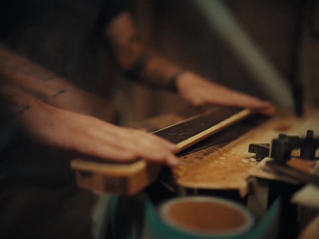 A craftsman with tattooed arms meticulously sands the wooden fretboard of a guitar neck in a dusty workshop