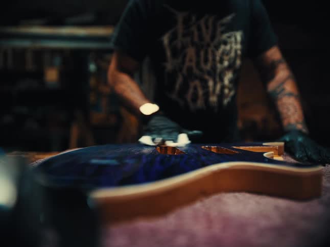 A tattooed artisan meticulously polishes the deep blue body of a custom electric guitar in a dimly lit workshop