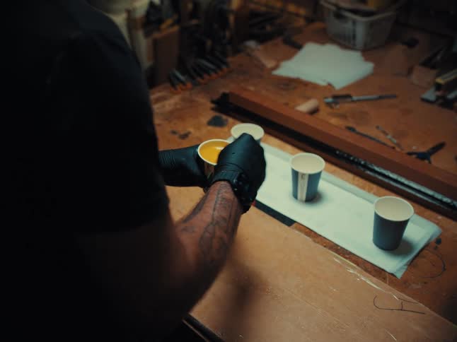 A craftsman wearing black gloves mixes yellow epoxy resin in a paper cup at a workbench in his workshop