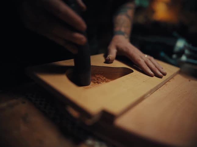 A craftsman with tattooed arms meticulously sands a piece of wood using a rotary tool in a workshop