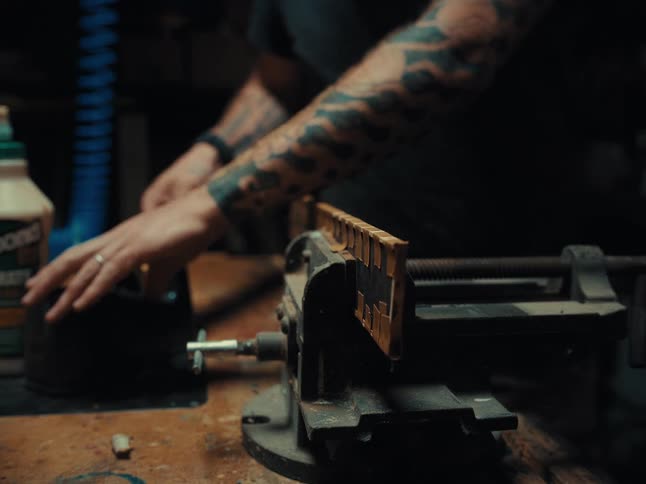 A tattooed craftsman meticulously tapes a wooden piece clamped in a vise within his dimly lit, dusty workshop