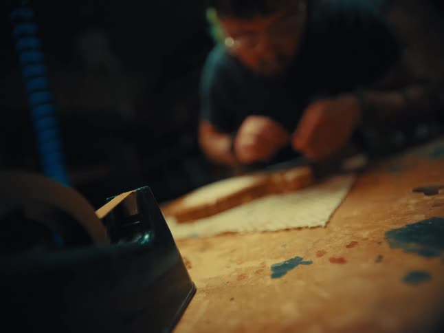 A tattooed artisan uses a tape dispenser to carefully prepare a project on a wooden workbench in a dimly lit workshop