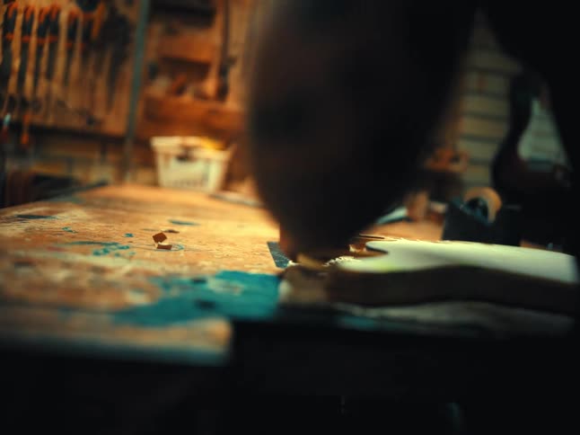 A craftsman with tattooed arms uses a scraper and heat gun while working on a wooden guitar body in their workshop