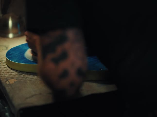 A craftsman with tattooed arms meticulously sands a vibrant blue electric guitar body with a power sander in a dimly lit workshop