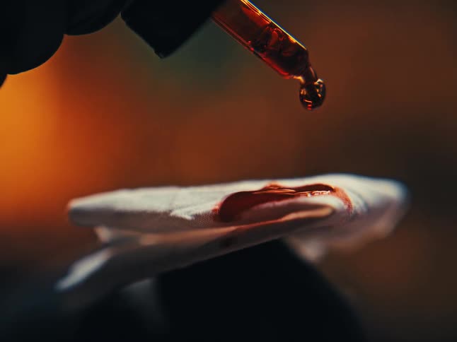 An intense macro shot shows a reddish-brown liquid being dropped from a pipette onto a white cotton pad in slow motion