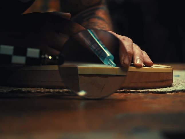 A craftsman carefully applies glue with a syringe to the body of a wooden guitar, viewed through a magnifying glass