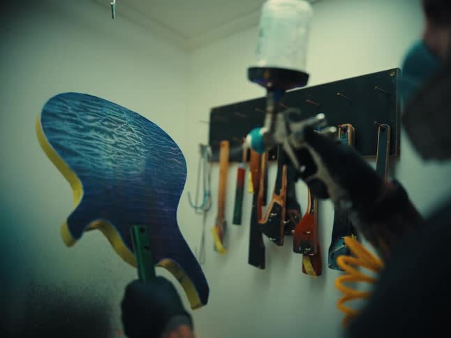 A luthier carefully spray-paints a clear varnish onto a custom blue electric guitar body in slow motion