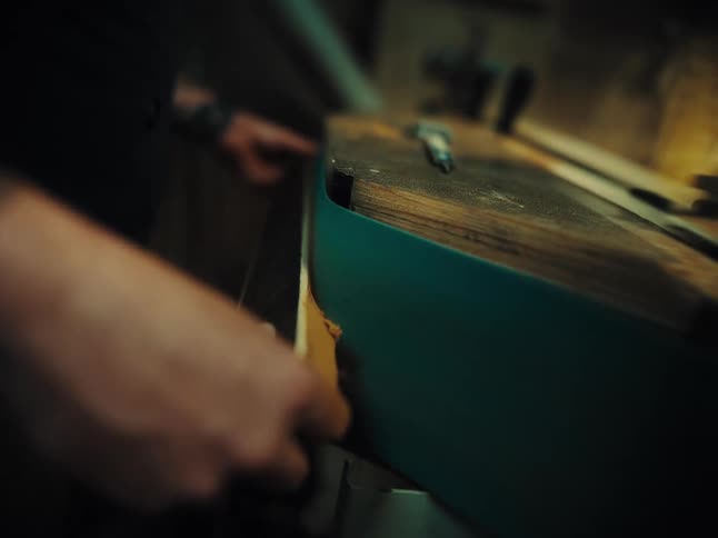 A close-up, shallow focus shot shows a craftsperson sanding the edge of a wooden board on a belt sander in a moody workshop