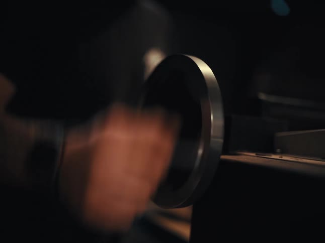 A man rapidly turns a large metal crank on a machine in a dark, moody setting