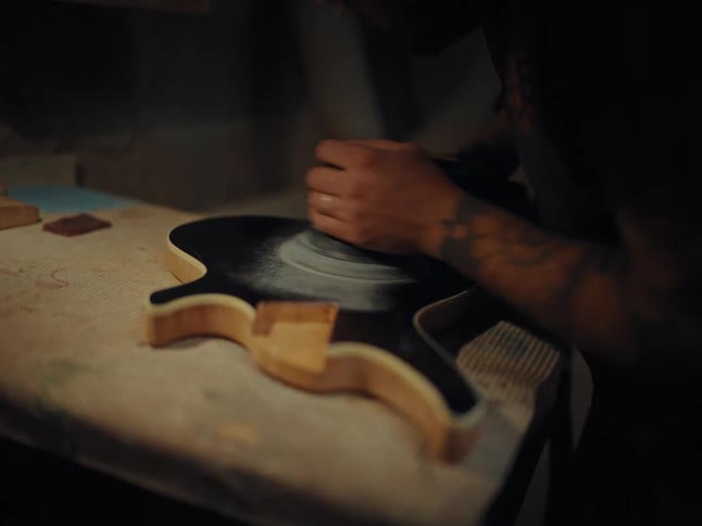 A craftsman with tattoos uses an orbital sander to smooth the black body of an electric guitar in a dimly lit workshop