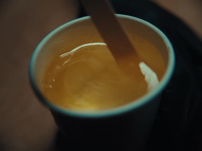 Close-up of a wooden stir stick mixing a warm, golden-colored beverage in a dark paper cup