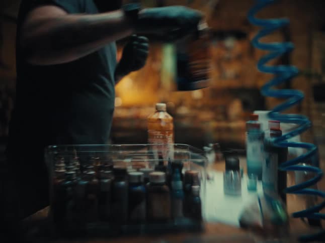 A tattooed artist wearing gloves opens and prepares various paints and chemicals in his workshop
