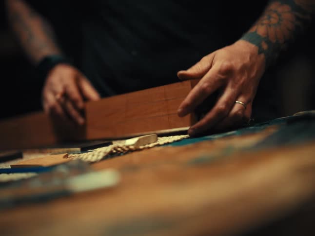 A tattooed craftsman carefully measures and draws lines with a pencil on a piece of wood in a dimly lit workshop
