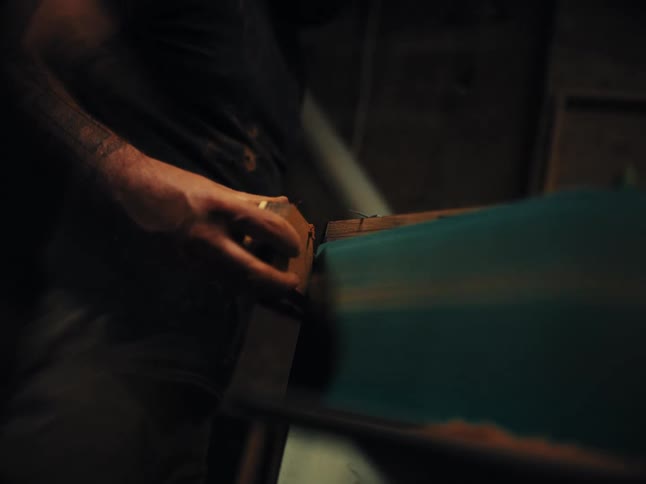 A tattooed craftsman carefully sands a piece of wood on a belt sander in a dimly lit workshop