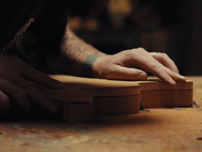 A tattooed luthier carefully shapes the wooden body of a guitar on a router table in his workshop