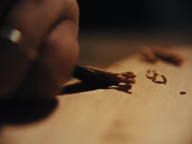 A craftsman meticulously carves a piece of wood with a sharp chisel, creating delicate shavings in a warmly lit workshop
