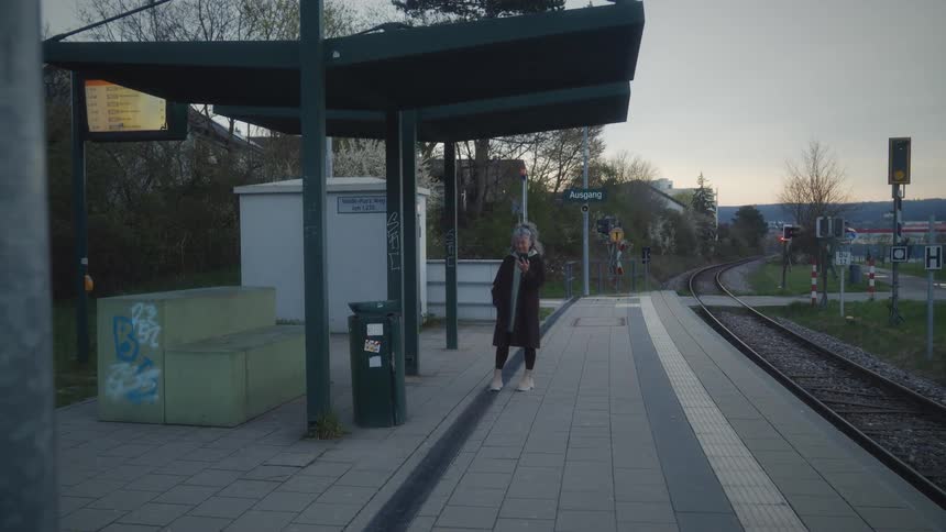 A woman with grey hair stands alone on a quiet train station platform in the evening, talking on her phone