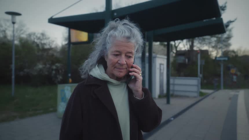A close-up shot of a smiling senior woman with grey hair on a phone call zooms out to reveal her waiting alone at a tram station at dusk
