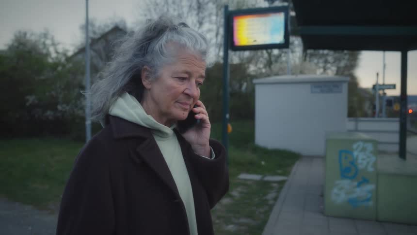 A senior woman with long gray hair answers her phone and has a conversation while walking to a tram stop at dusk in slow motion