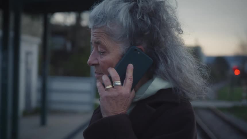 An older woman with gray hair has a serious conversation on her smartphone while waiting at a train station at dusk
