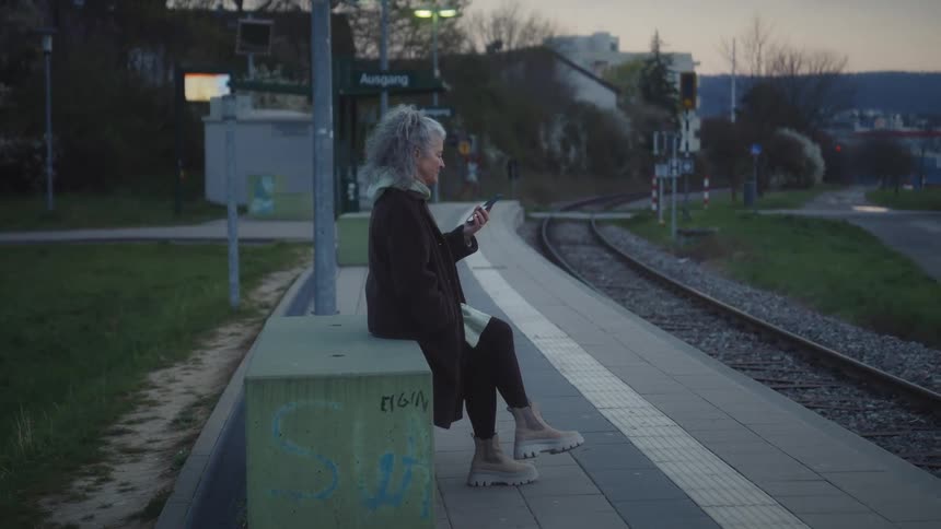 A stylish older woman with curly gray hair finishes a phone call and waits patiently on a quiet train platform at dusk