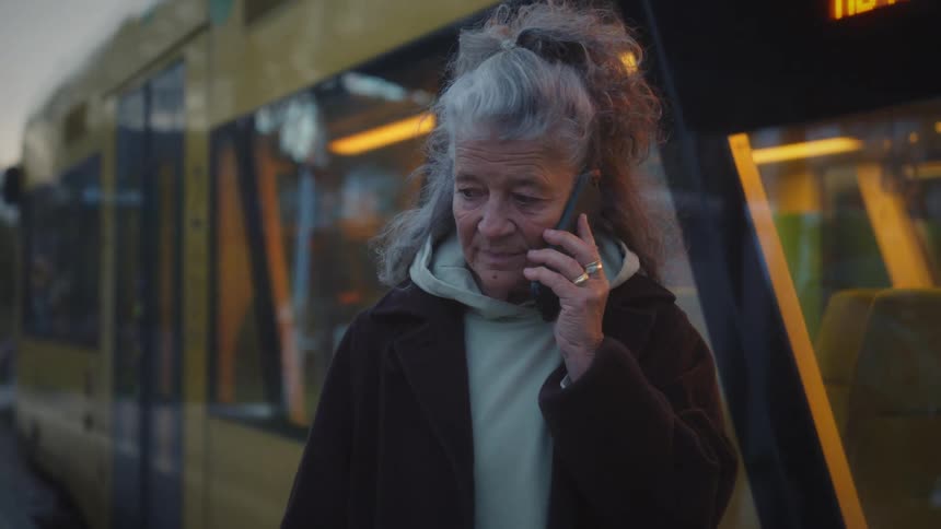 An elderly woman with grey hair talks on her phone at a train station as a tram pulls in during the evening