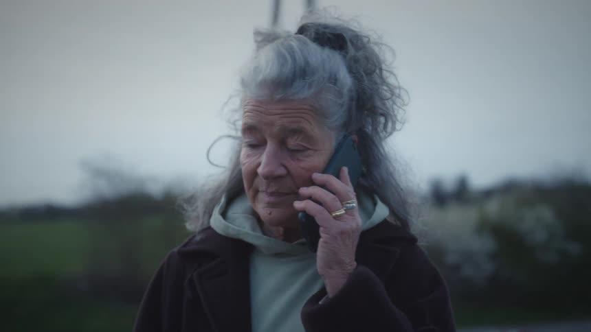 A senior woman with curly grey hair has a conversation on her smartphone while walking along a quiet train platform at twilight