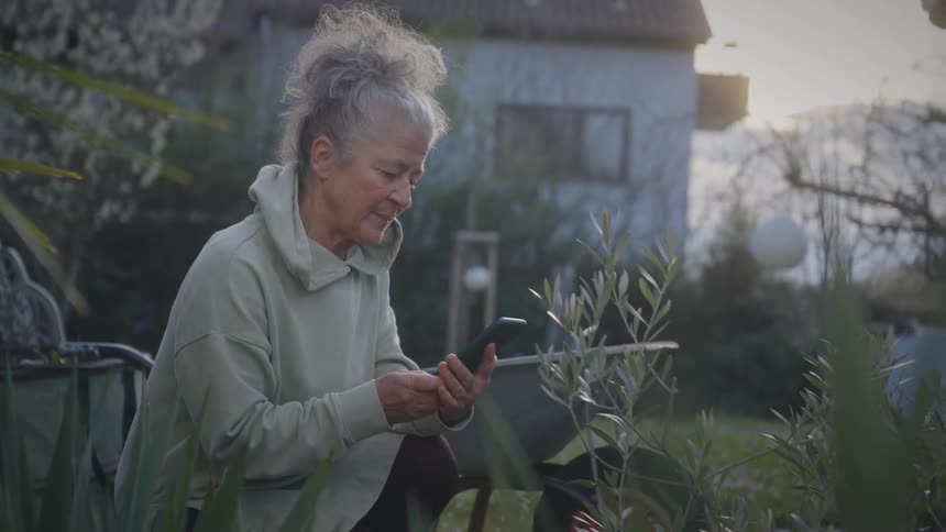A senior woman takes a phone call while working in her garden and then uses her smartphone to check on her plants