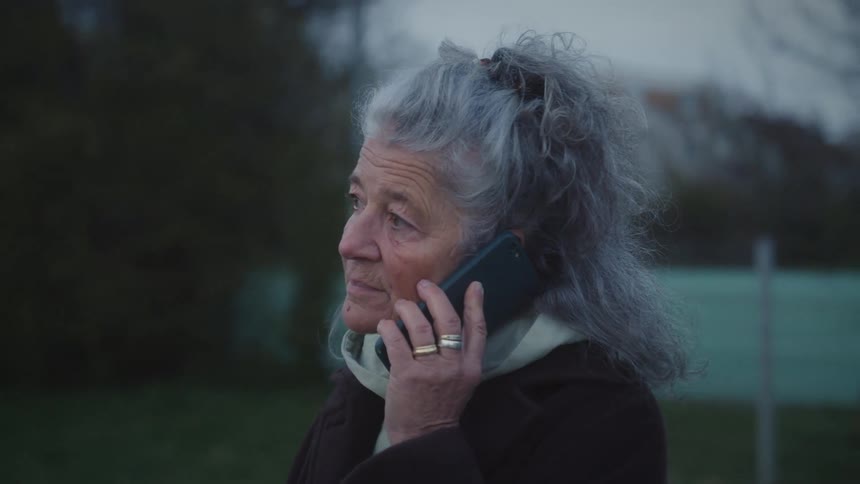 A thoughtful senior woman with long grey hair talks on her smartphone while standing outside in the evening light
